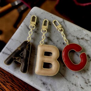 Può includere: Tre portachiavi a forma di lettera A, B e C. La lettera A è marrone con un motivo a monogramma scuro, la lettera B è beige e la lettera C è rossa. Ogni lettera è imbottita, cucita e fissata a una catenella in metallo color oro.