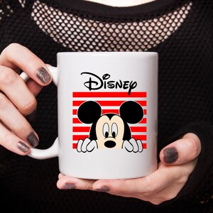 May include: White ceramic mug featuring Mickey Mouse peeking out from behind a red and white striped rectangle. The Disney logo is above the graphic. The mug is held by a person.