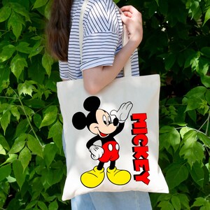 May include: A cream-colored tote bag featuring a cartoon image of Mickey Mouse waving. Mickey is depicted in his classic outfit with red shorts, yellow shoes, and white gloves. The word "MICKEY" is printed in red, vertical letters.