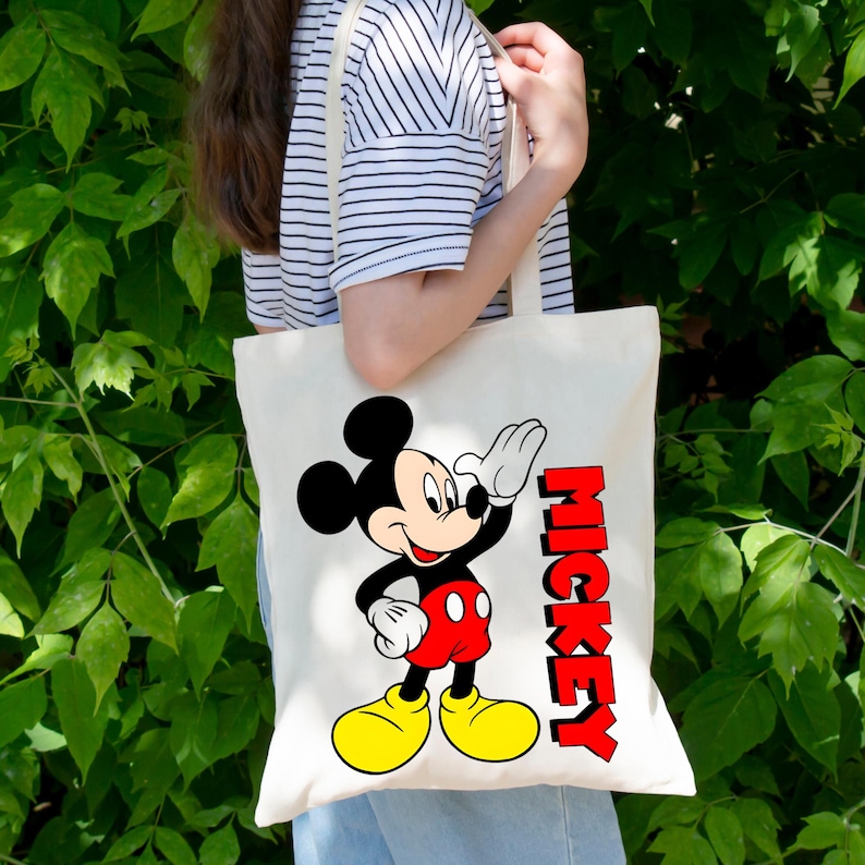 May include: A cream-colored tote bag featuring a cartoon image of Mickey Mouse waving. Mickey is depicted in his classic outfit with red shorts, yellow shoes, and white gloves. The word "MICKEY" is printed in red, vertical letters.