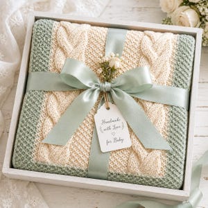May include: A square, cream-colored knitted blanket with a cable knit pattern and sage green borders, presented in a white wooden box. It is tied with a sage green satin ribbon, a small sprig of dried flowers, and a tag reading Handmade with Love for Baby.