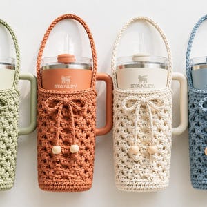 May include: Four Stanley tumblers in a row, each in a different color: green, orange, cream, and blue. Each tumbler is in a matching crocheted holder with a handle and a bow. The Stanley logo is visible on each tumbler.