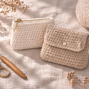 Könnte beinhalten: Zwei gehäkelte Beutel, einer cremefarben mit goldenem Reißverschluss und der andere beige mit Knopfverschluss, liegen auf einer neutralen Oberfläche. Ein Häkelhaken und eine goldene Schere liegen neben den Beuteln, mit einem Wollknäuel im Hintergrund.