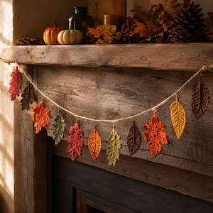 Op de afbeelding: Een decoratieve slinger met een reeks gehaakte bladeren in diverse herfstkleuren, waaronder rood, oranje, geel, bruin en groen, allemaal aan elkaar geregen aan een dun, lichtgekleurd touw voor een hangende presentatie.