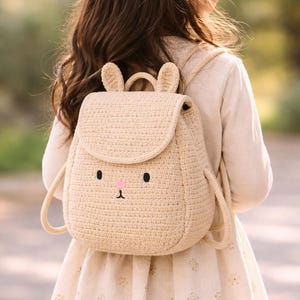 May include: A beige crochet backpack with a bunny face design. The backpack features a flap closure, adjustable straps, and a top handle. The bunny face includes black eyes, a pink nose, and small ears. The backpack is designed for children.