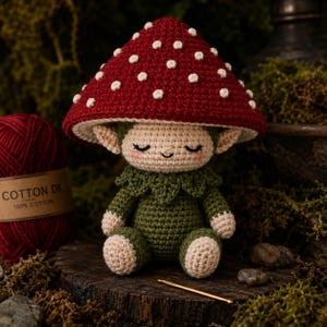 May include: A crocheted doll featuring a large, deep red mushroom cap with white spots, a beige face with closed eyes and a small smile, and a dark green body with leaf-like collar details. The doll is crafted from yarn and has pointed ears.