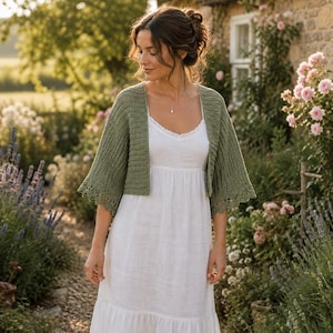 Peut inclure: Un boléro en crochet vert sauge avec une texture côtelée et un bord en dentelle festonnée sur les manches, porté sur une robe blanche sans manches simple avec une jupe froncée et une fine bordure en dentelle le long de l'encolure.