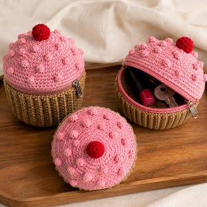May include: Three crochet cupcake-shaped pouches with tan ribbed bases and pink tops featuring small raised bobble textures and a single red pom-pom on each. One pouch is open, revealing a lipstick and a metal key inside the zippered compartment.