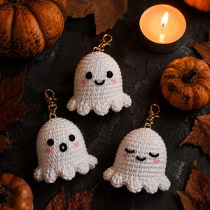 May include: Three white crochet ghost keychains with gold-colored clasps, each with a different facial expression. The ghosts have black eyes and pink cheeks. The background includes pumpkins, leaves, and a lit candle, creating a Halloween theme.
