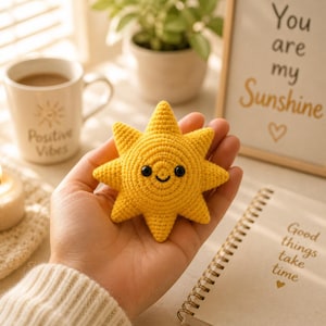 May include: A handmade yellow crochet sun with a smiling face and black eyes. The sun is held in a hand, with a mug that says "Positive Vibes" and a framed print that says "You are my Sunshine" in the background. A notebook with the text "Good things take time" is also visible.