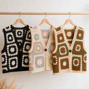 Può includere: Tre gilet all'uncinetto in nero, pastello e verde oliva sono appesi a grucce di legno. I gilet presentano un motivo a punto nonna quadrato con chiusure a bottoni. Il gilet nero ha un motivo color crema, mentre il gilet pastello ha un mix di colori.
