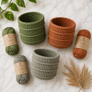 Könnte beinhalten: Drei gehäkelte Körbe in Grün, Orange und Grau mit passenden Garnknäueln. Die Garnetiketten tragen die Aufschrift "#4 Worsted Cotton" und listen Farben wie "Cactus Green" und "Terracotta" auf. Die Körbe sind zylindrisch und handgefertigt.