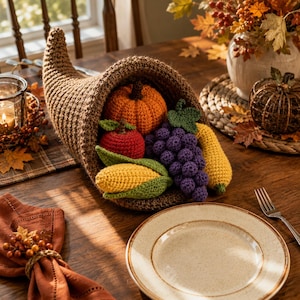 May include: A brown crocheted cornucopia filled with various crocheted produce including an orange pumpkin, a red apple, a bunch of purple grapes, a yellow ear of corn with green husks, and a yellow squash, all arranged as a centerpiece.