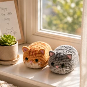 Op de afbeelding: Twee gehaakte kattenspeeltjes, één oranje en één grijs, staan op een vensterbank. Beide hebben roze oren, zwarte ogen en witte snorharen. Een ingelijste print met de tekst "Enjoy the little things" en een kleine vetplant zijn ook zichtbaar.
