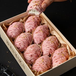 May include: A box of pink pastries with a layered, textured appearance, possibly meringue or a similar confection. Each pastry is topped with gold leaf flakes. The box is beige with the brand name "HOLYMARN" printed on the side.