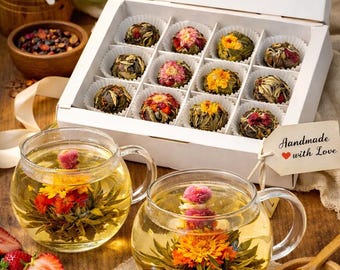 May include: A white box with a clear window contains 12 blooming tea balls. Two glass teacups with handles hold tea with blooming flowers. The scene includes a wooden honey dipper, strawberries, and a tag that says "Handmade with Love."