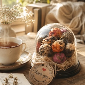 May include: A glass cloche filled with dried flowers sits on a wooden surface, next to a teacup and saucer. A small tag tied to the cloche reads "thank you." The scene is warmly lit, suggesting a cozy, inviting atmosphere. A vase of white flowers is also visible.