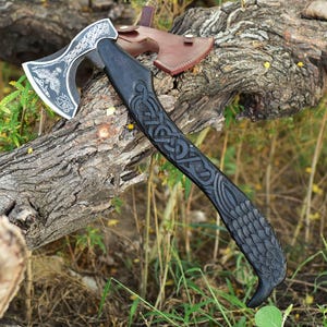 May include: A hand axe with a dark wooden handle featuring intricate carved knotwork and feather patterns. The metal blade displays an etched design of a bird and decorative scrollwork. A brown leather sheath is attached to the axe head.