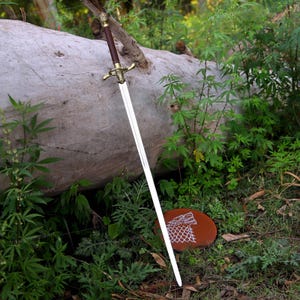 May include: A long sword with a dark brown leather-wrapped handle, a metallic gold-toned crossguard, and a straight steel blade. A circular brown leather plaque featuring a white embossed wolf head design rests on the ground near the blade.