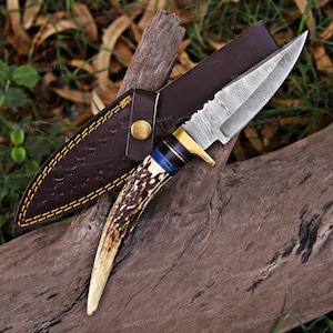 May include: A handcrafted hunting knife with a patterned steel blade and an antler handle with blue accents. The knife is paired with a brown leather sheath with decorative stitching and a brass button clasp. The knife is resting on a piece of wood.