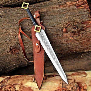 May include: A double-edged dagger with a pointed silver blade featuring an etched fan-like pattern. The handle is black with a square brass pommel and guard. It is accompanied by a brown leather sheath with a strap and metal snap fasteners.