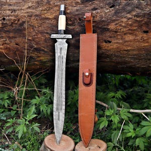 May include: A Damascus steel sword with a bone handle and a brown leather sheath. The blade has a detailed pattern, and the sheath has a belt loop and a snap closure. The sword is displayed on a wooden surface.