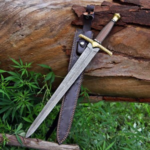 May include: A double-edged sword with a patterned steel blade, a gold-coloured crossguard, and a wrapped handle, accompanied by a dark brown leather scabbard featuring a snap closure and decorative stitching along the edges.