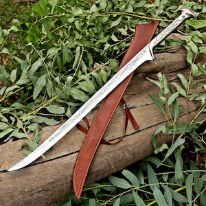 May include: A silver-coloured metal sword with a curved blade and an ornate handle featuring intricate engraved patterns. The sword is accompanied by a brown leather scabbard with visible stitching and thin leather straps for attachment.