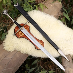 May include: A silver-toned metal sword with a black leather-wrapped grip, a crossguard, and a pommel. The blade features engraved runic text. A black scabbard with silver-toned metal fittings lies alongside the sword on a decorative wooden plaque.