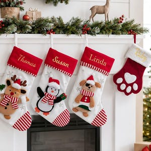 May include: Four Christmas stockings hanging from a white mantel. Three white stockings feature red cuffs with the names Thomas, Susan, and Linda embroidered in gold, plus plush reindeer, penguin, and dog appliqués. One red paw-shaped stocking.