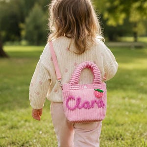 May include: A light pink corduroy handbag featuring a textured surface, two top handles, and a detachable shoulder strap. The front is decorated with the name Clara in dark pink cursive lettering and a small red strawberry applique with green leaves.