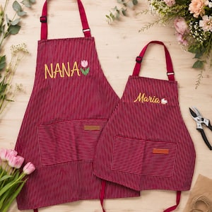 May include: Two matching burgundy aprons with thin white vertical stripes. The larger apron features the embroidered name NANA with a pink tulip, while the smaller apron displays the name Maria with a small cupcake icon. Both have front pockets.