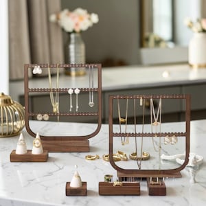 May include: Two wooden jewelry organizers with various necklaces and earrings displayed. The organizers are dark brown and have a scalloped top edge. Several small ring holders are also present, along with a few rings and bracelets. The jewelry is gold and silver.