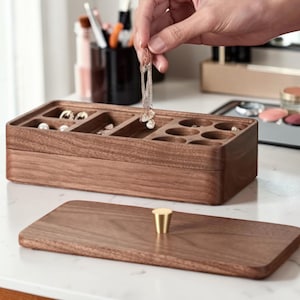 Pode incluir: Uma caixa de joias de madeira com formato retangular e tampa removível. A caixa é dividida em compartimentos de vários tamanhos para guardar joias. Uma mão segura um par de brincos acima da caixa. A caixa é feita de madeira marrom escura.