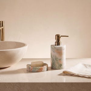 May include: A bathroom set featuring a cylindrical marble soap dispenser with a brass pump, a matching round marble soap dish holding a bar of soap, and a beige cloth napkin, positioned next to a large stone basin with a brass faucet.