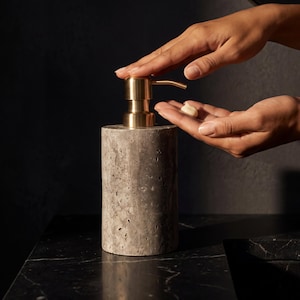 Peut inclure: Un distributeur de savon cylindrique en pierre grise poreuse avec un mécanisme de pompe en métal doré brossé. Le distributeur présente une surface naturelle texturée avec de petites indentations sur tout le corps en pierre, surmonté d'un embout métallique.