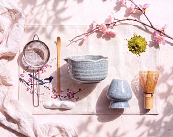 May include: A matcha tea set featuring a blue speckled ceramic bowl with a scalloped rim, a matching blue whisk holder, a bamboo whisk, a bamboo scoop, a metal mesh strainer, and a small white ceramic rest, arranged with green matcha powder.