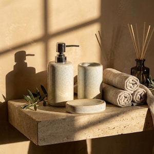May include: A bathroom accessory set featuring a ceramic soap dispenser with a silver pump, a matching tumbler, and a soap dish. The set is accompanied by three rolled beige towels, a glass reed diffuser with wooden sticks, and a small olive branch.