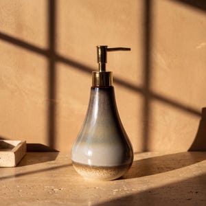 May include: A teardrop-shaped ceramic soap dispenser with a glossy finish that transitions from a dark charcoal top to a light gray middle and a speckled beige base, topped with a metallic gold-toned pump mechanism for dispensing liquid soap.