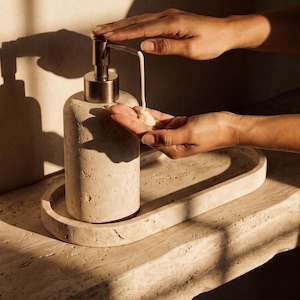 May include: A cylindrical beige stone soap dispenser with a metallic pump mechanism, paired with a matching oval stone tray. The dispenser has a porous, textured surface and is shown dispensing a small amount of cream-coloured liquid soap.