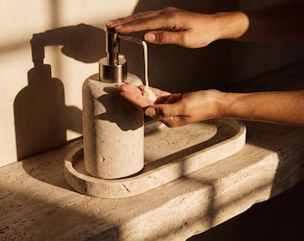May include: A cylindrical beige stone soap dispenser with a metallic pump mechanism, paired with a matching oval stone tray. The textured, porous surface of the stone features natural variations in color and tone, creating a cohesive set.
