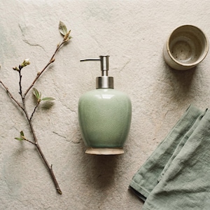 May include: A sage green ceramic soap dispenser with a metallic pump mechanism, accompanied by a small beige ceramic cup, a folded sage green linen cloth, and a thin brown branch with small buds, all arranged on a textured stone surface.