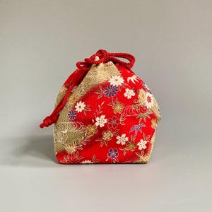 May include: A small drawstring pouch featuring a vibrant red and gold fabric with a detailed floral pattern. The bag is cinched at the top with a thick red cord, showcasing white, purple, and gold flower motifs across the textured material.