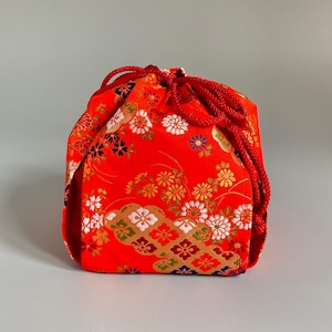 May include: A bright red drawstring pouch featuring a repeating pattern of white, gold, and blue floral motifs. The bag is cinched at the top with matching red braided cords, creating a gathered closure. The fabric displays intricate woven details.