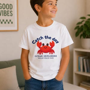 Könnte beinhalten: Ein weißes kurzärmeliges T-Shirt mit einer roten Cartoon-Krabbe in der Mitte. Über der Krabbe steht der Text Catch the day in Blau. Unter der Krabbe steht der Text OCEAN EXPLORERS MALIBU BEACH CLUB in Blau. Das T-Shirt wird mit einer blauen Jeans getragen.