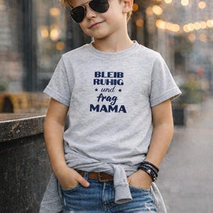 Könnte beinhalten: Ein Kind trägt ein graues T-Shirt mit dem marineblauen Text "BLEIB RUHIG * und * frag MAMA". Es trägt auch eine blaue Jeans, eine Sonnenbrille und ein Armband. Ein graues Sweatshirt ist um die Taille gebunden.