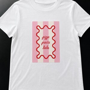 Könnte beinhalten: Weißes T-Shirt mit einem rechteckigen Grafikdesign mit rosa und weißen Streifen. Die Grafik hat einen roten, wellenförmigen Rand und die Worte "pizza pasta liebe". Das T-Shirt hat kurze Ärmel und einen Rundhalsausschnitt.