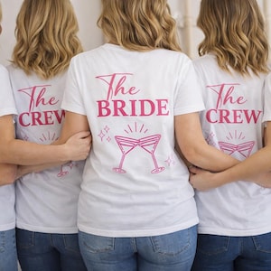 Könnte beinhalten: Weiße T-Shirts mit rosa Text und Grafiken. Die Shirts tragen auf der Rückseite die Aufschrift "The Crew" und "The Bride". Die Grafik zeigt zwei gekreuzte Cocktailgläser. Die Personen tragen blaue Jeans.