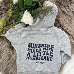 Könnte beinhalten: Grauer Kapuzenpullover mit dem marineblauen Text "SUNSHINE MIXED WITH A LITTLE HURRICANE" und "est.2024". Der Hoodie hat eine Kapuze und ist aus weichem Material. Weiße Blumen und Grünzeug sind im Hintergrund.