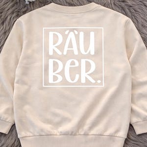 Könnte beinhalten: Ein beigefarbener Langarm-Pullover mit einem weißen rechteckigen Rahmen auf der Rückseite, der den Text RÄUBER enthält. Das Kleidungsstück hat einen Rundhalsausschnitt sowie gerippte Bündchen an Ärmeln und Saum aus einfarbigem Stoff.